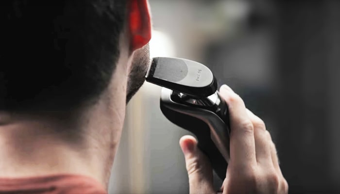 The back of a dark-haired man’s head who is bringing an electric shaver close to his cheek.