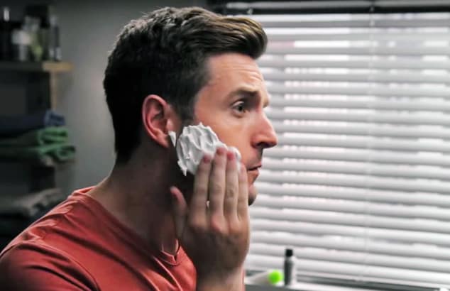 Profile of a brown-haired man rubbing white shaving foam onto his face as he looks to the side.