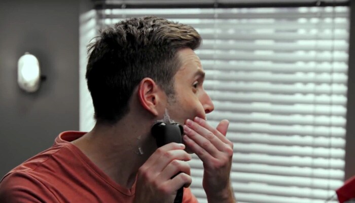 A man using his hands to push his face to the side to shave his cheek with an electric razor.