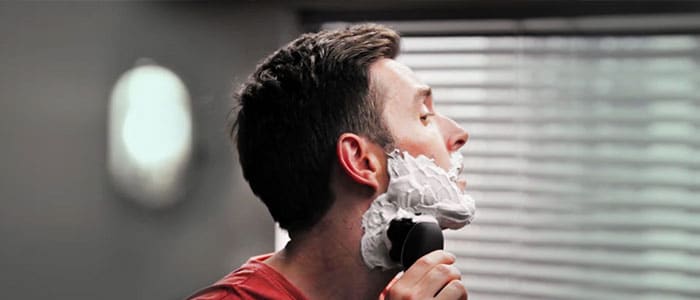 A man with shaving foam on his face as he shaves with an electric razor.