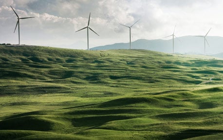 A photograph of a green hillside with wind turbines spinning to create renewable energy in the background