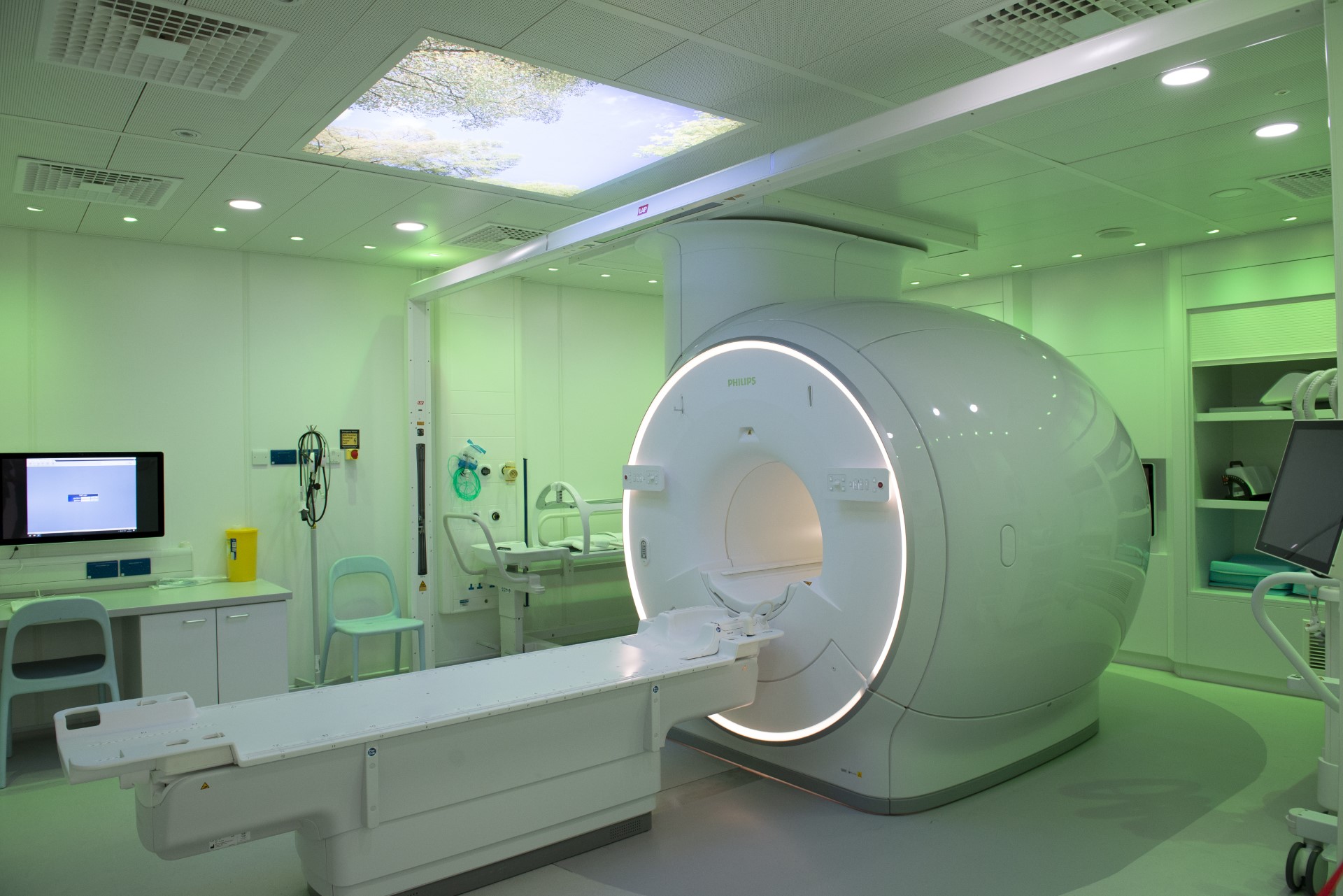 Image shows the new Philips MRI simulator situated in Leeds Hospital. There is a large MRI machine in white surrounded by green lighting in a hospital room.