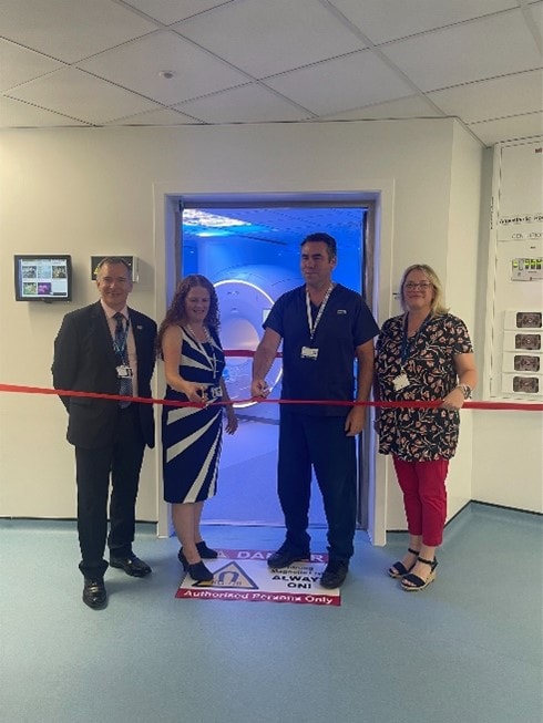 This photo shows four NHS staff cutting a red ribbon in front of a room lit up in blue which holds the Philips MRI scanner.