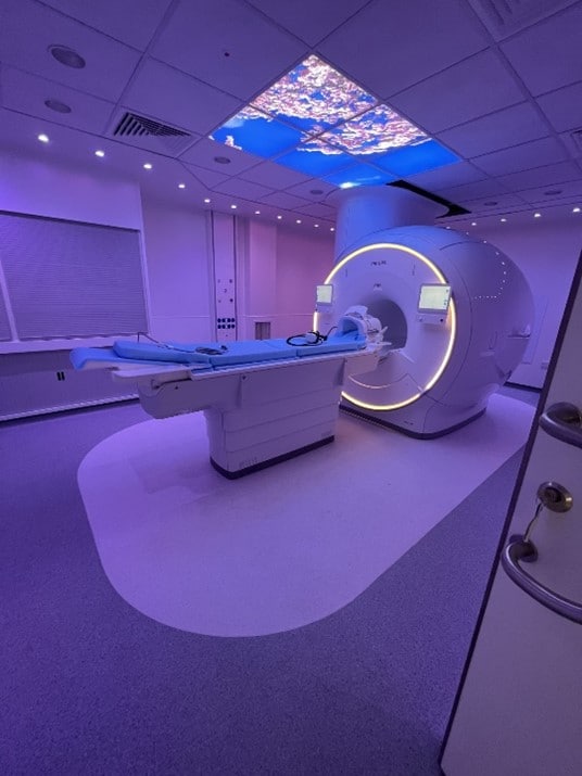 This photo shows the new Philips MRI system complete with Philips Ambient Experience situated in a hospital room. The room is lit up in purple and has an image of blossom trees projected on the ceiling above the MRI.