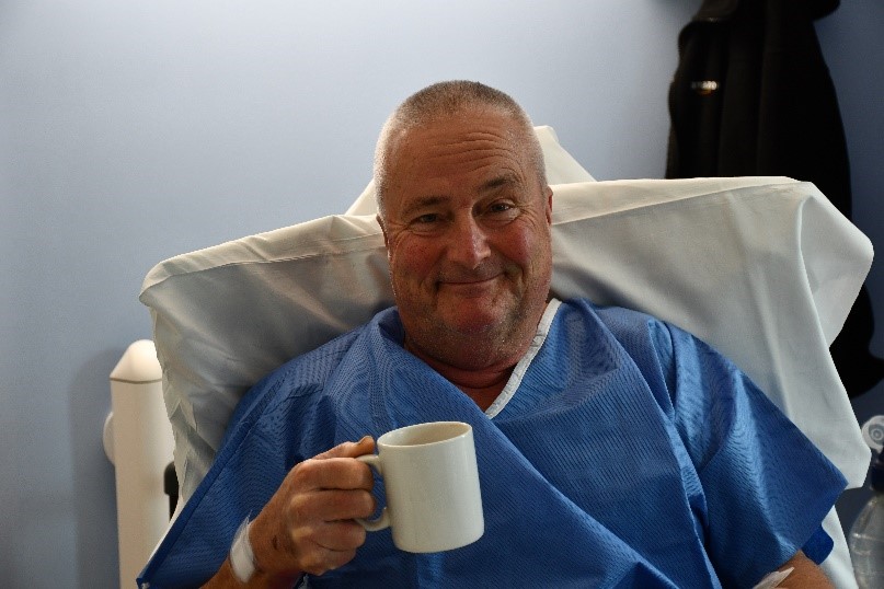 Michael Bronze, sitting in the day ward at Royal Papworth Hospital after his procedure in a blue surgical gown. Michael is holding a cup of tea and smiling.